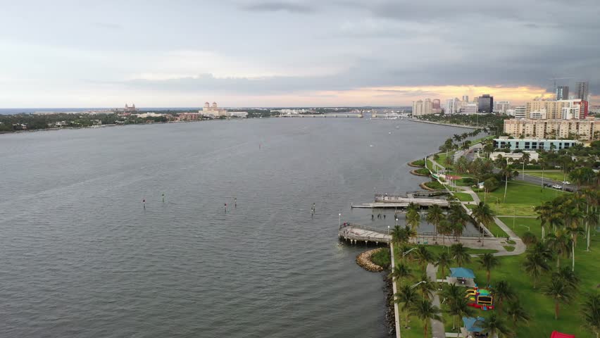 Aerial view of the Intracoastal Waterway with waterfront parks, palm trees, and the city skyline under a cloudy sunset sky in West Palm Beach, Florida, USA.