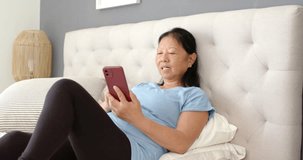 Senior Asian woman in blue tee holding maroon phone tapping after alert smiling by tufted headboard. Bedroom, bed, pillows, lamp, device, cozy, relaxed - Powered by Shutterstock - Get 15% off with code: PIKWIZARD15