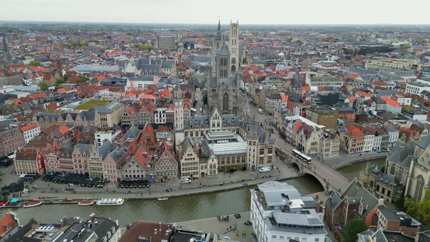 Aerial drone shot of the historic center of Ghent, Belgium. Featuring the Leie river with tour boats, St. Michael’s Bridge, and the Gothic architecture of St. Nicholas Church.