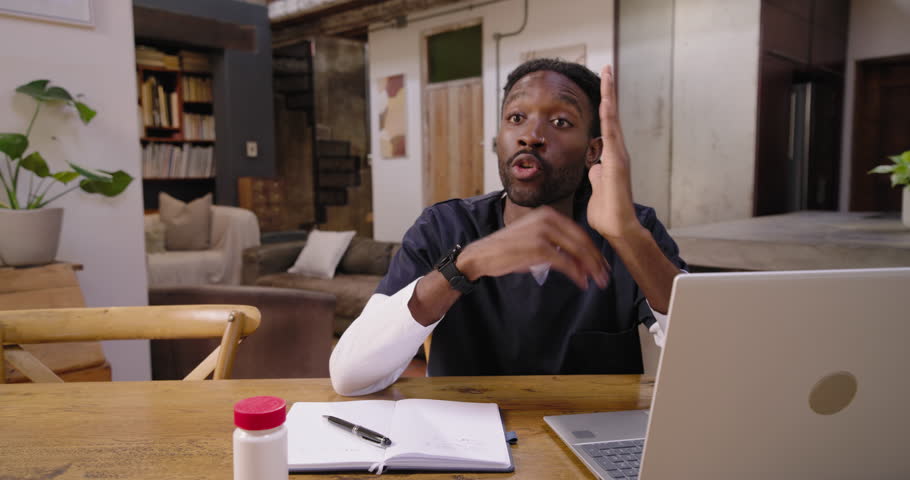 Man in scrubs sitting at table, aligning to DSLR appearing, glancing at laptop, showing pill bottle. Presenter, camera, tripod, loft, home, gestures, tutorial