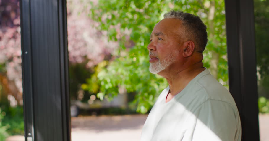 African American senior man standing on porch, dappled sun on face, scanning yard and smiling. Elder, gentleman, doorway, path, trees, shrubs, sunlight