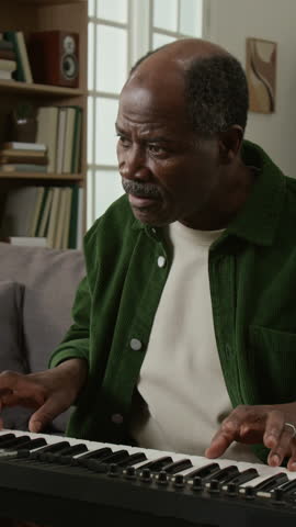 Vertical shot of happy mature Black man learning to play piano at home and turning looking at camera with smile, reading sheet music