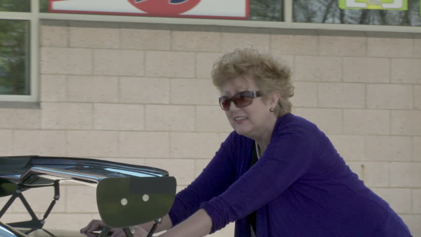 Mature woman pushes a car up to the pumps at a gas station while man looks on. 