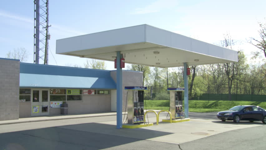 Car drives up to pumps at unbranded gas station.  Wide shot with camera moving