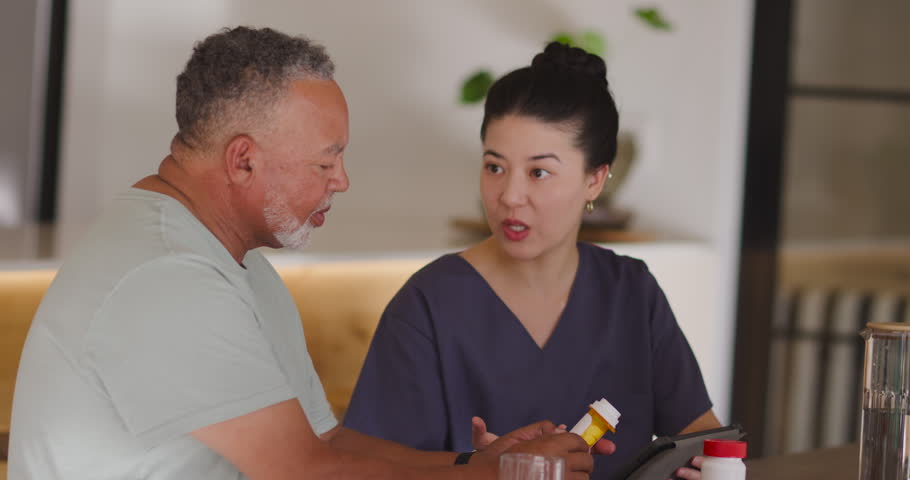 Asian caregiver holding yellow bottle and Senior man clarifying dosage using tablet at home table. Medication, pill, device, screen, kitchen, consultation, guidance