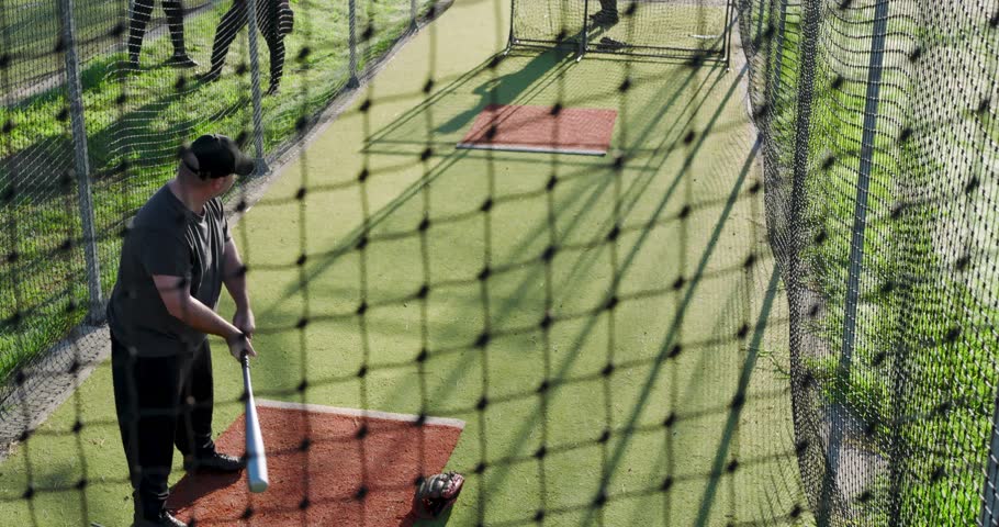 Mature adult male batter picking up bat in batting cage swinging at pitching machine for practice. Athletic, teamwork, training, sport, coaching, outdoor, determined