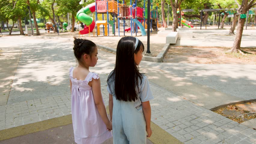 Two young girls stand holding hands in a sunny park playground during a static shot