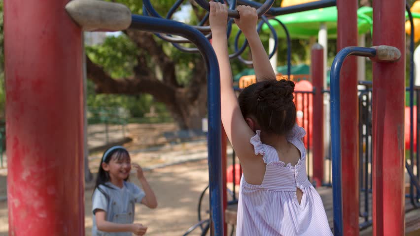 Young Thai girl hangs from monkey bars before dropping down to play with her friend