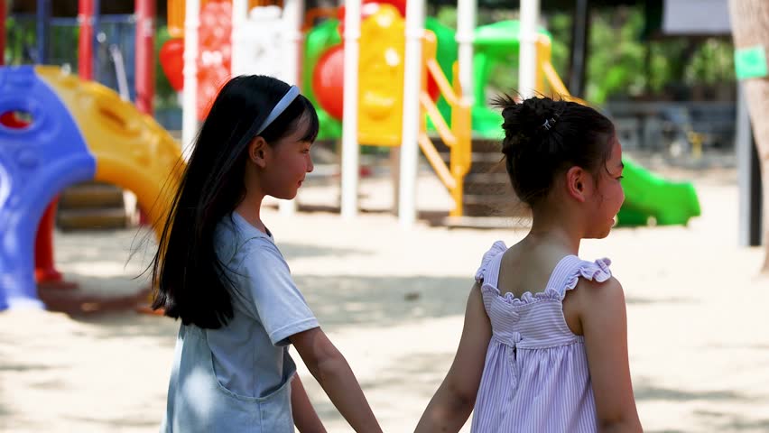 Two young girls hold hands while walking past colorful slides in a bright outdoor playground