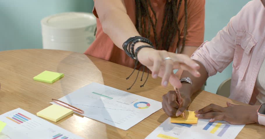 Left woman reaching at table is starting; right woman pressing notes and sorting charts for review. Collaboration, teamwork, meeting, office, data, graphs, planning