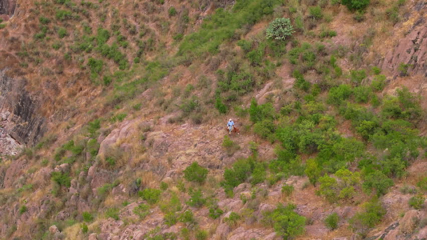 Man on horse carefully makes his way through challenging rocky cliff edge at height. Aerial drone footage captures rider traversing steep mountainside with green shrubs, boulders, and a stream below.