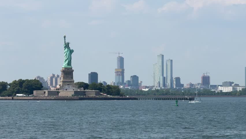 The Statue of Liberty stands on Liberty Island with New York Harbor waters and distant city skyline behind it. Iconic American landmark represents freedom, travel, and tourism under clear blue sky.