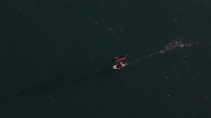 Aerial view of a person wing foiling across dark rippling water with a white wake.
