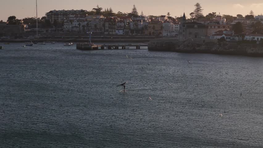 Aerial view of a person wing foiling on the calm sea near the historic waterfront and pier during golden hour in Cascais, Lisbon, Portugal.
