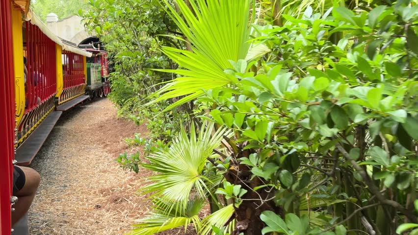 A colorful small train moves along a narrow track into a stone tunnel surrounded by dense green plants. The charming amusement-style railway creates a playful outdoor travel scene.