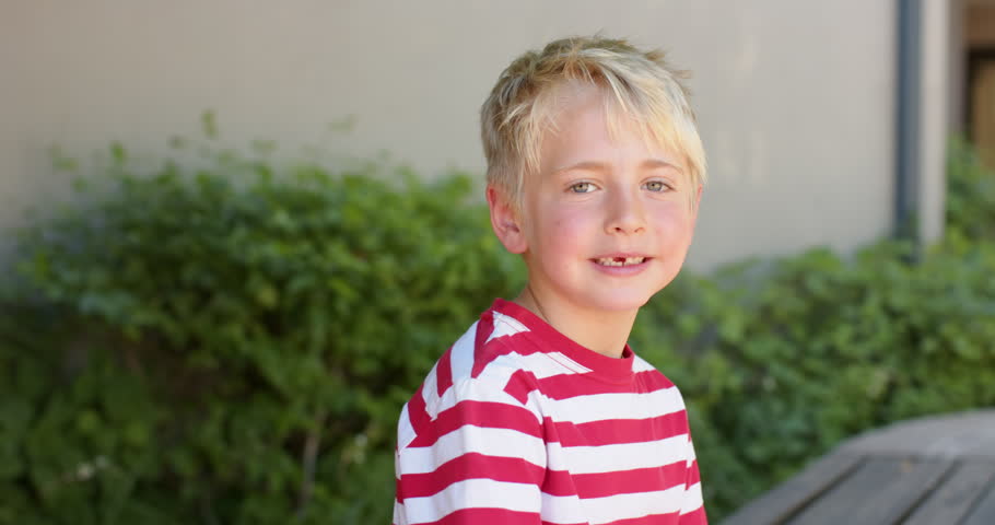 School-age boy sitting on wooden bench, shifting torso and smiling with camera framing tightening. Child, outdoor, courtyard, greenery, seat, striped, sunny