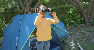 Clip starting with child raising binoculars by blue tent, scanning trees near green backpack. Camping, outdoors, kid, adventure, canopy, sunshine, nature - Powered by Shutterstock - Get 15% off with code: PIKWIZARD15