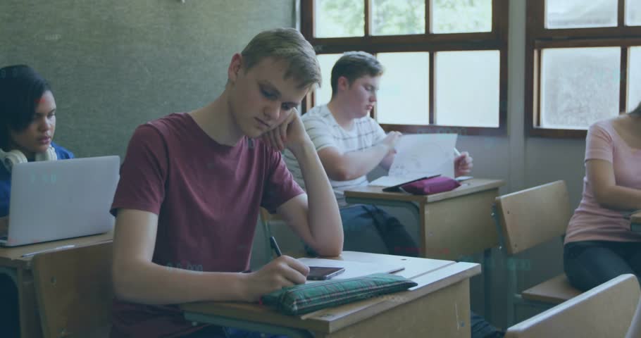 Tapping smartphone, teen male student wearing maroon tee raising head toward camera at front desk. Classroom, laptop, headphones, daylight, window, learning, group