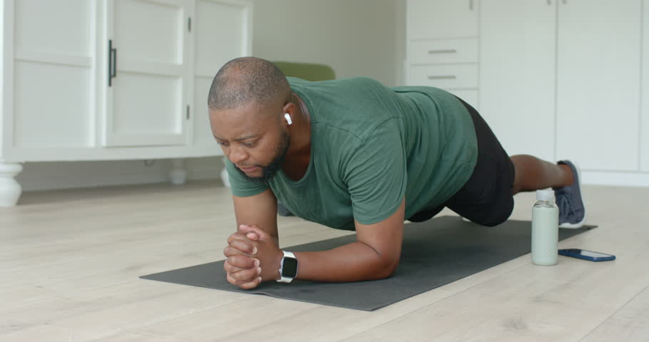 African American man holding plank on mat at home, hearing earbuds, ending hold and laughing. Apartment, core, fitness, training, stability, minimalist, bright