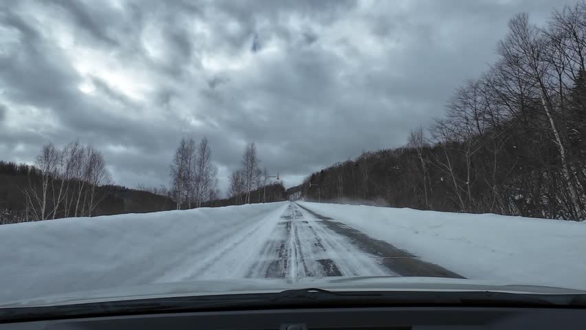 POV driving through a narrow snow covered road with high snow banks forming a corridor in Hokkaido winter landscape under cloudy sky