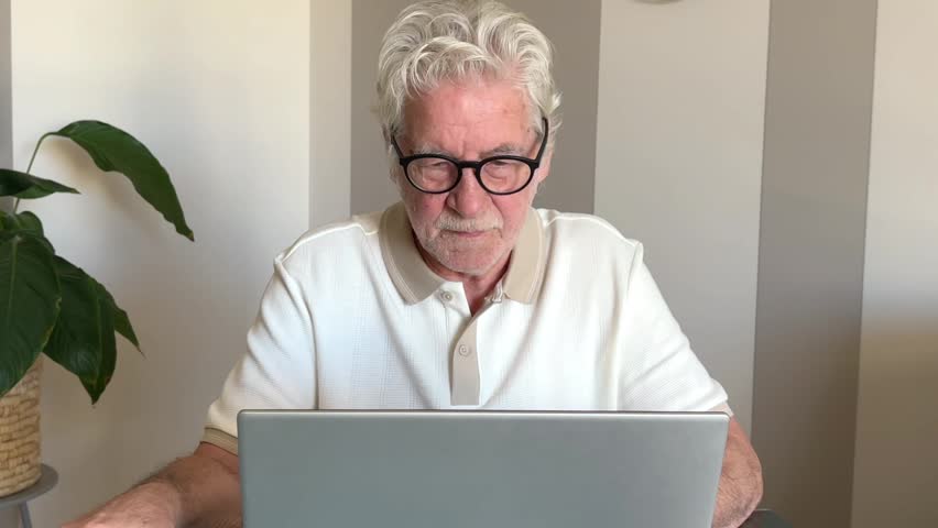 An elderly man happily smiles and laughs, engaging with his laptop. He expresses joy and connection while using technology at home, potentially during a video call or browsing content.