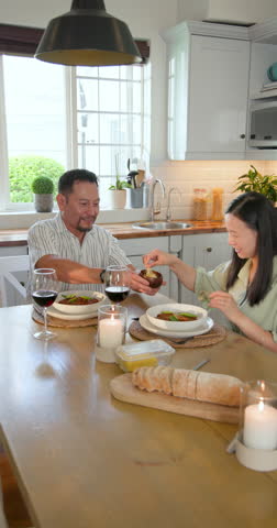 Vertical video: Asian couple reaching for dressing and buttering bread for meal at kitchen table. Romantic, intimacy, dining, healthy, lifestyle, rustic, vertical video