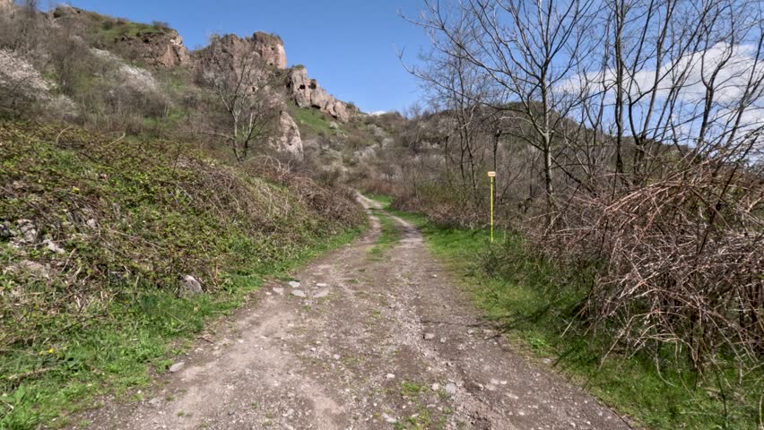 People walk along a dirt path surrounded by hills and trees. The sun shines bright as nature blooms in spring. The trail invites exploration and adventure.