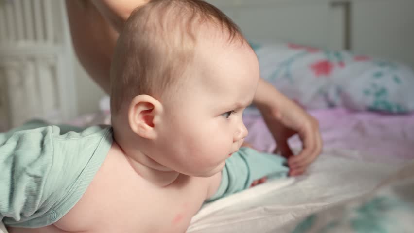 A mother dresses her baby girl after bathing on a bed while the baby is playing and looking at her.