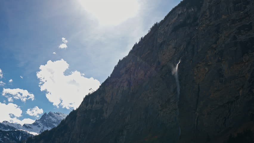 A dramatic low-angle view of a towering sheer rock cliff with a slender waterfall cascading down its face and a mountain cable lift visible against a bright cloudy sky in a deep Swiss alpine valley.