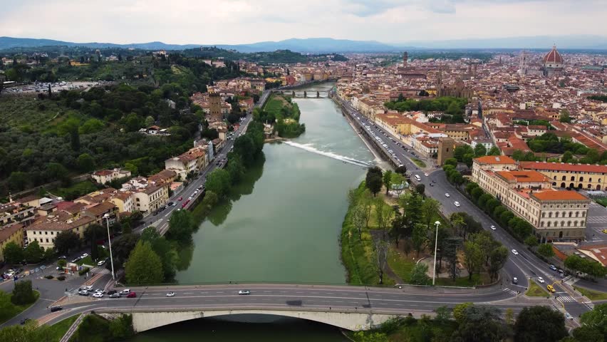 Drone shot over the Arno river embankments in Florence, Italy. Historic city center with the Cathedral dome and Santa Croce basilica visible across the rooftops, Tuscan hills in the background.