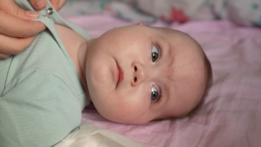A mother dresses her baby girl after bathing on a bed while the baby is playing and looking at her.