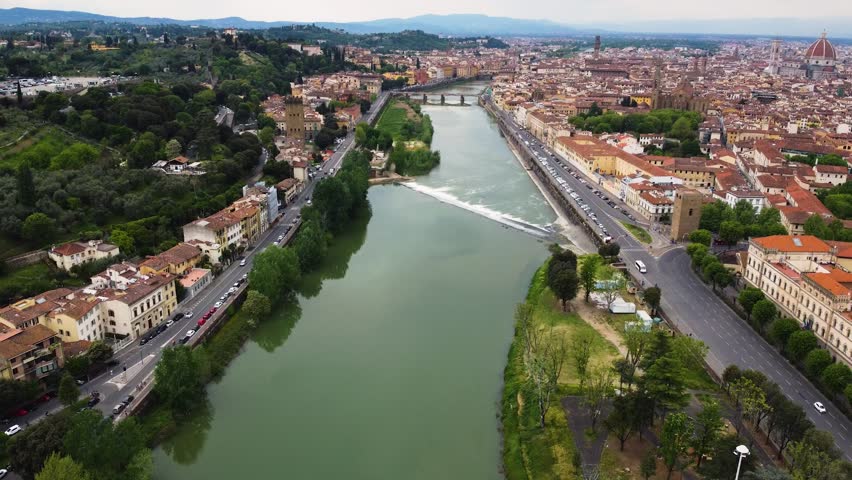 Aerial drone shot along the Arno river through Florence, Italy. Historic bridges, city embankments and the Cathedral of Santa Maria del Fiore dome visible across the full skyline.