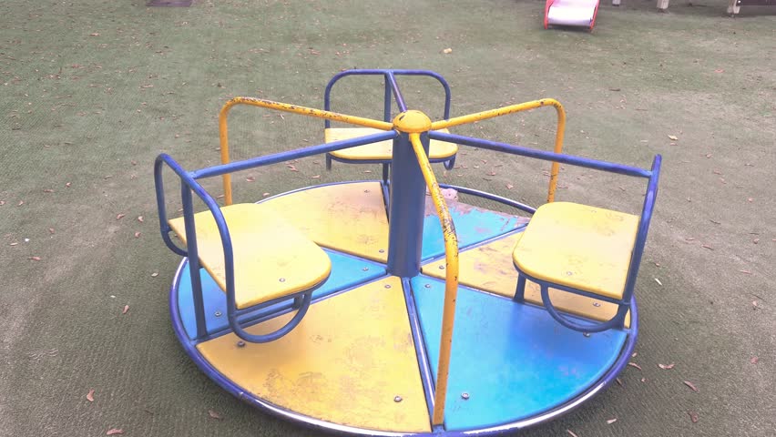 A yellow and blue playground carousel rotating slowly in an outdoor park. Park equipment, leisure time, childhood activities.