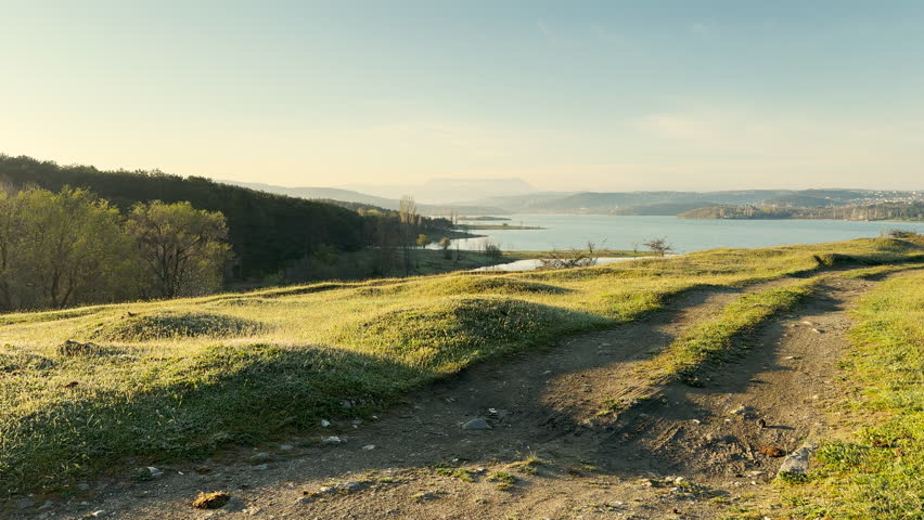 Grassy hills roll gently toward a wide blue lake during a quiet morning. This vast landscape feels peaceful and free. The soft sunlight on the field creates a sense of pure relaxation.