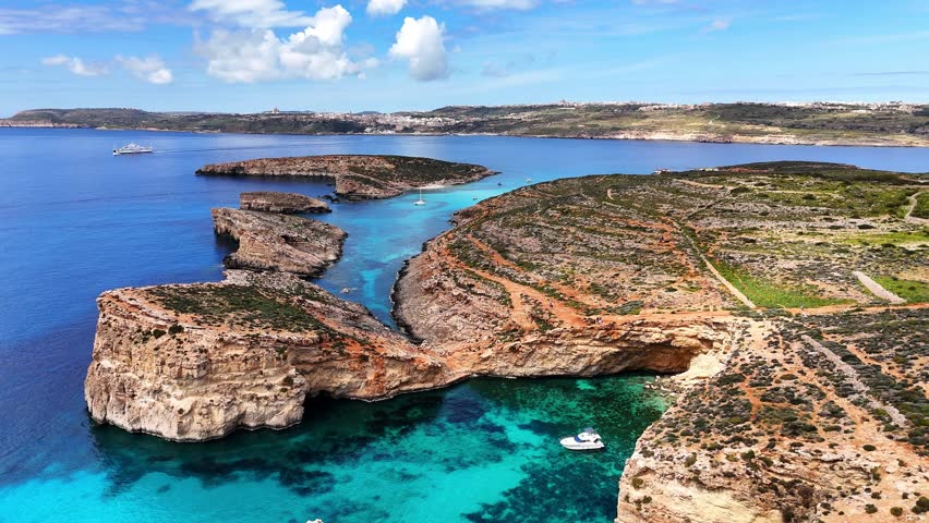 Aerial view of Blue Lagoon in Comino, Malta, showing a rocky bay, small islets, anchored boats and clear turquoise sea under sunny daylight.