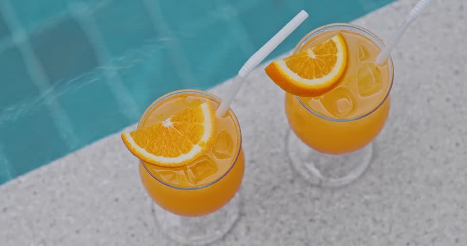 Orange juice glasses with fresh citrus slices sitting on swimming pool edge, turquoise resort water background, summer vacation lifestyle and travel advertisement