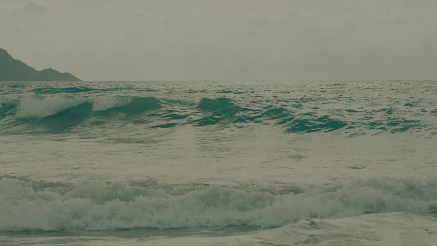 Clouds cover the sky as waves roll onto the sandy beach. The ocean foams and splashes against rocks. Mountains stand in the background. Water flows back into the sea.