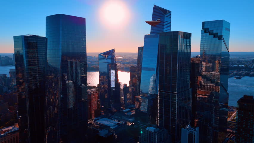 Sunset over Manhattan skyline featuring modern glass skyscraper architecture. High angle shot of tall buildings with a bright sun disk reflecting off glass facades in the evening.