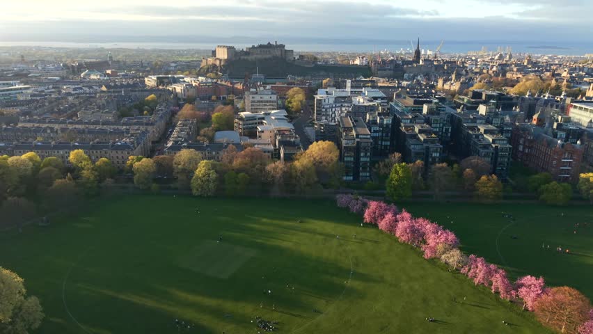 Aerial view of The Meadows park with blooming cherry blossoms and the historic Edinburgh Castle overlooking the city skyline in Edinburgh, Scotland, United Kingdom.
