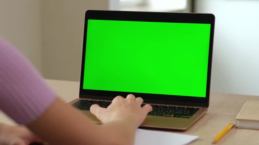 Back view of teen girl typing on keyboard of laptop with green screen. Female person using chroma key computer blank mock up at home
