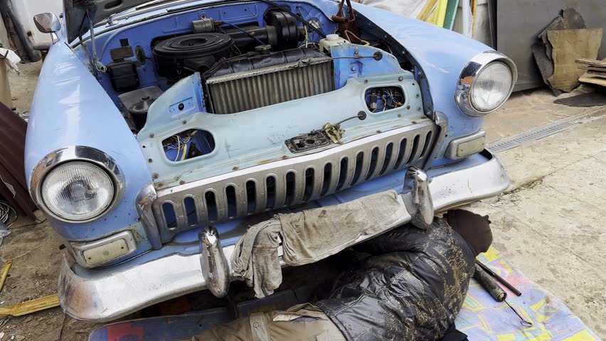 Male repairman lies under his vintage car at courtyard of house. Auto mechanic repairing his blue retro vehicle outdoor. Concept of automobile maintenance. Slow motion