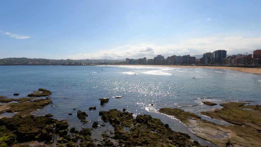 Stunning panoramic view of Playa San Lorenzo in Gijon, Asturias, Spain. Sparkling turquoise bay with rocky foreground, sandy beach, and urban skyline under bright blue sky.