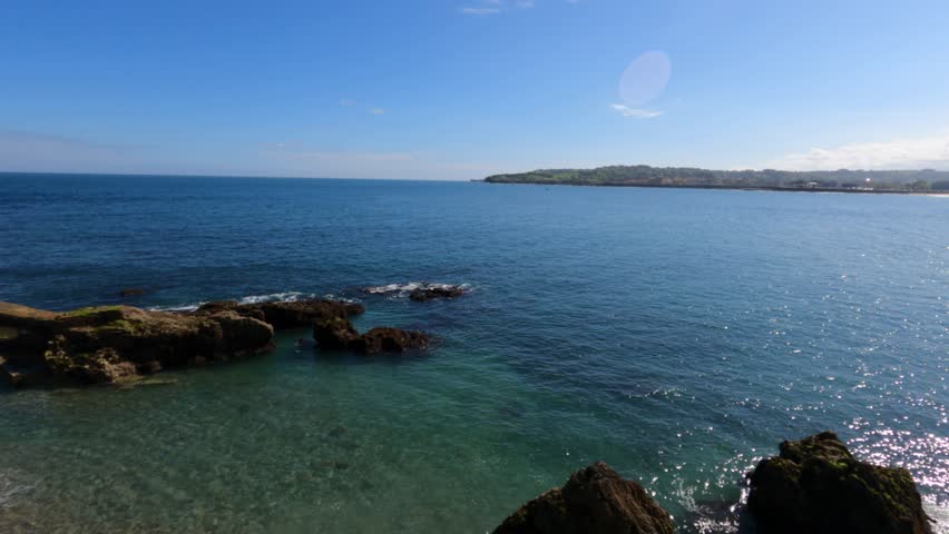 Stunning panoramic view of Playa San Lorenzo in Gijon, Spain, featuring turquoise waters, rugged rocky coastline, and a lush green headland under clear blue skies.