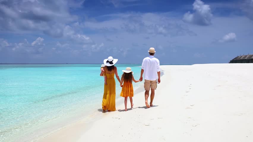 A family of four walks together on a tropical paradise beach with turquoise ocean and white sand during their vacation time in the Maldives islands