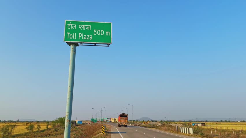 Wide highway scene featuring a green “Toll Plaza 500 m” sign in Hindi and English on a tall pole, with a heavy vehicle moving away in the background through open rural farmland under a clear blue sky.