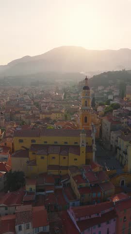 Aerial view of Menton, France, with the Basilique Saint Michel, pastel houses, red tiled roofs, promenade, docks, and groynes as the camera rises and pans down