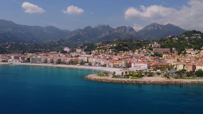 Aerial view of Menton, French Riviera, shows crescent pebble beach, promenade, curved stone breakwater, old quarter, Maritime Alps, gentle waves, people moving.