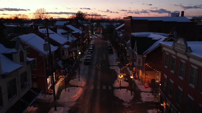 Historic small-town street at winter twilight with snow-covered rooftops, warm lantern lights and cozy two-story buildings. Charming and peaceful evening atmosphere. Aerial flyover shot.