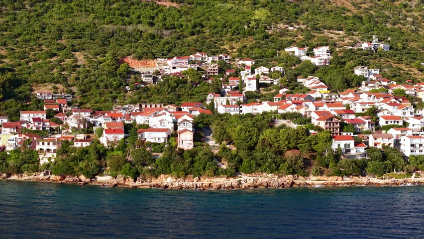 Aerial footage of a scenic coastal village in Croatia, Mediterranean houses with red roofs on a lush green hill overlooking the blue sea
