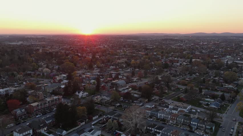 Aerial flyover residential neighborhood in York, Pennsylvania, USA at sunset, showing streets, rooftops and homes bathed in warm golden light with expansive urban landscape and horizon. Wide shot